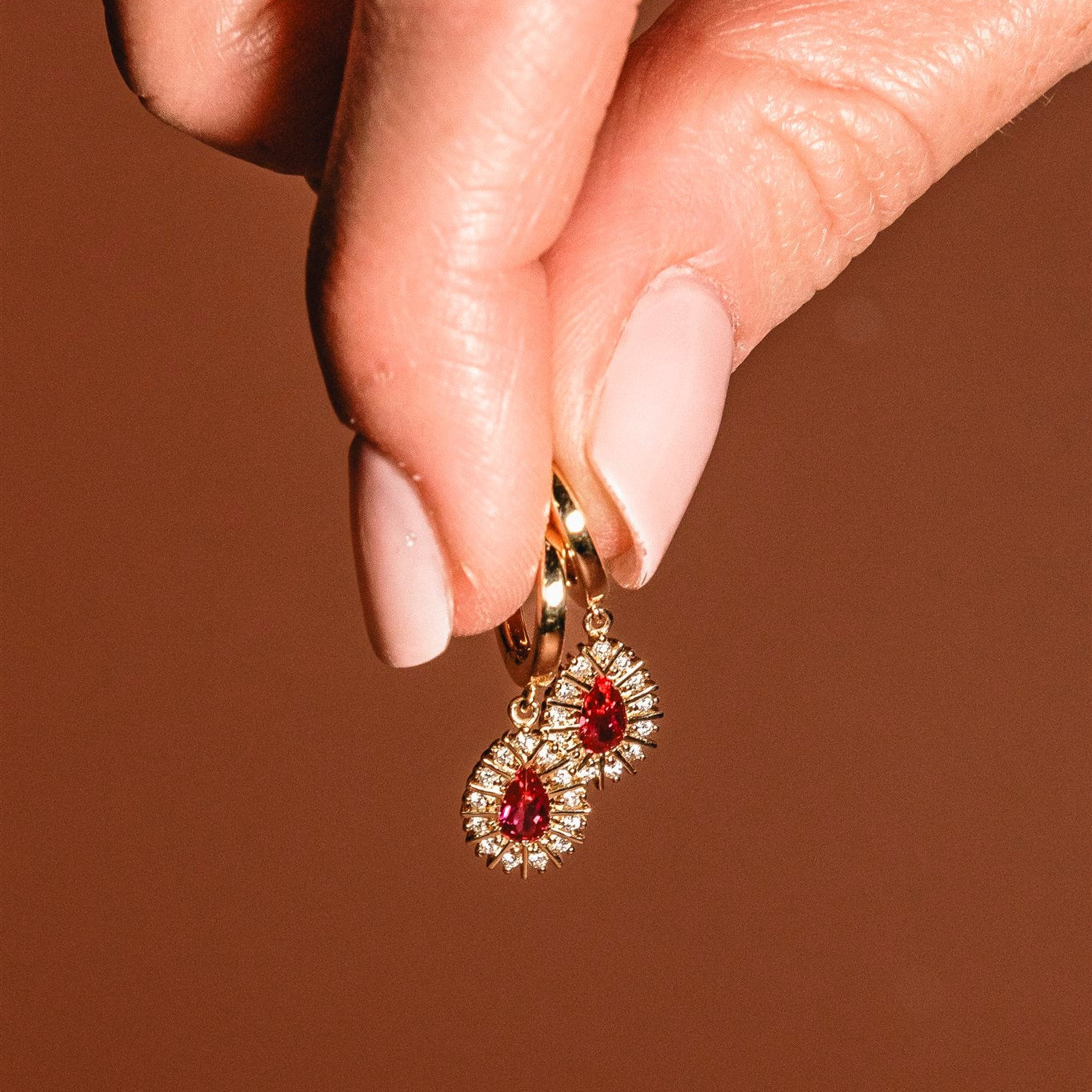 Gold earrings with red gemstones held by a hand against a brown background