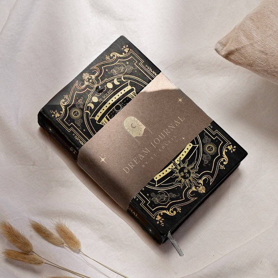 A vegan leather dream journal with gold foiling and a black obsidian crystal embedded on the cover, presented on a soft, light background with some plant elements.