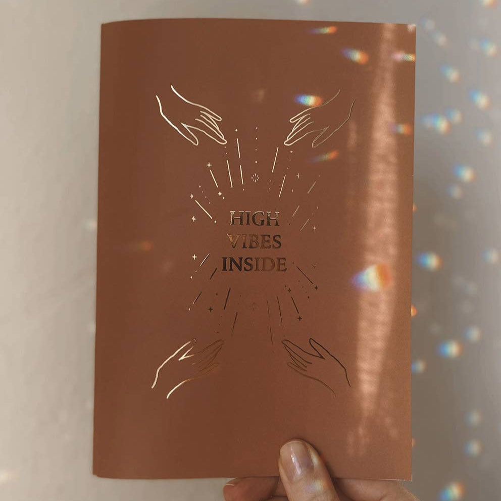 Brown notebook with 'High Vibes Inside' text and hand illustrations held by a person.