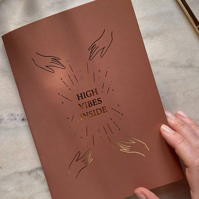 Brown book cover with gold text 'High Vibes Inside' and hand illustrations on a beige background