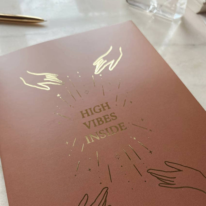 Brown book cover with gold text 'High Vibes Inside' and hand illustrations on a beige background