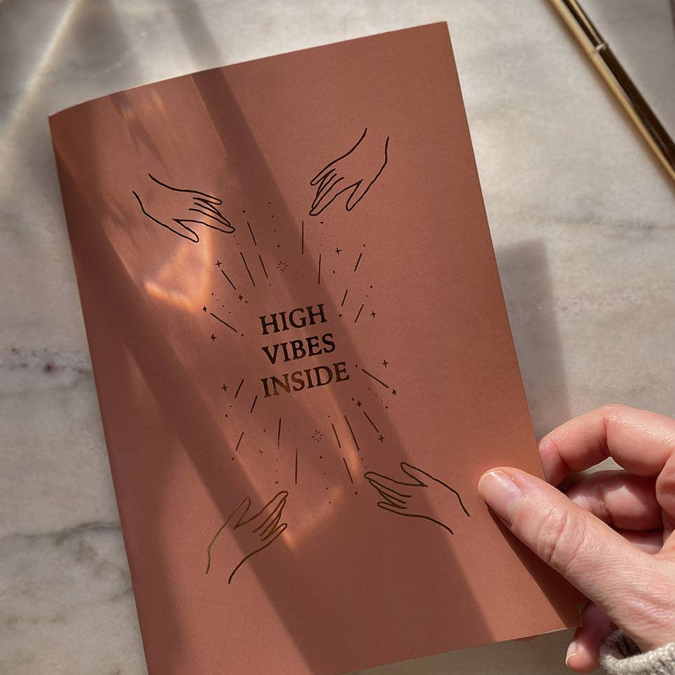 Brown notebook with 'High Vibes Inside' text and hand illustrations on a marble surface.