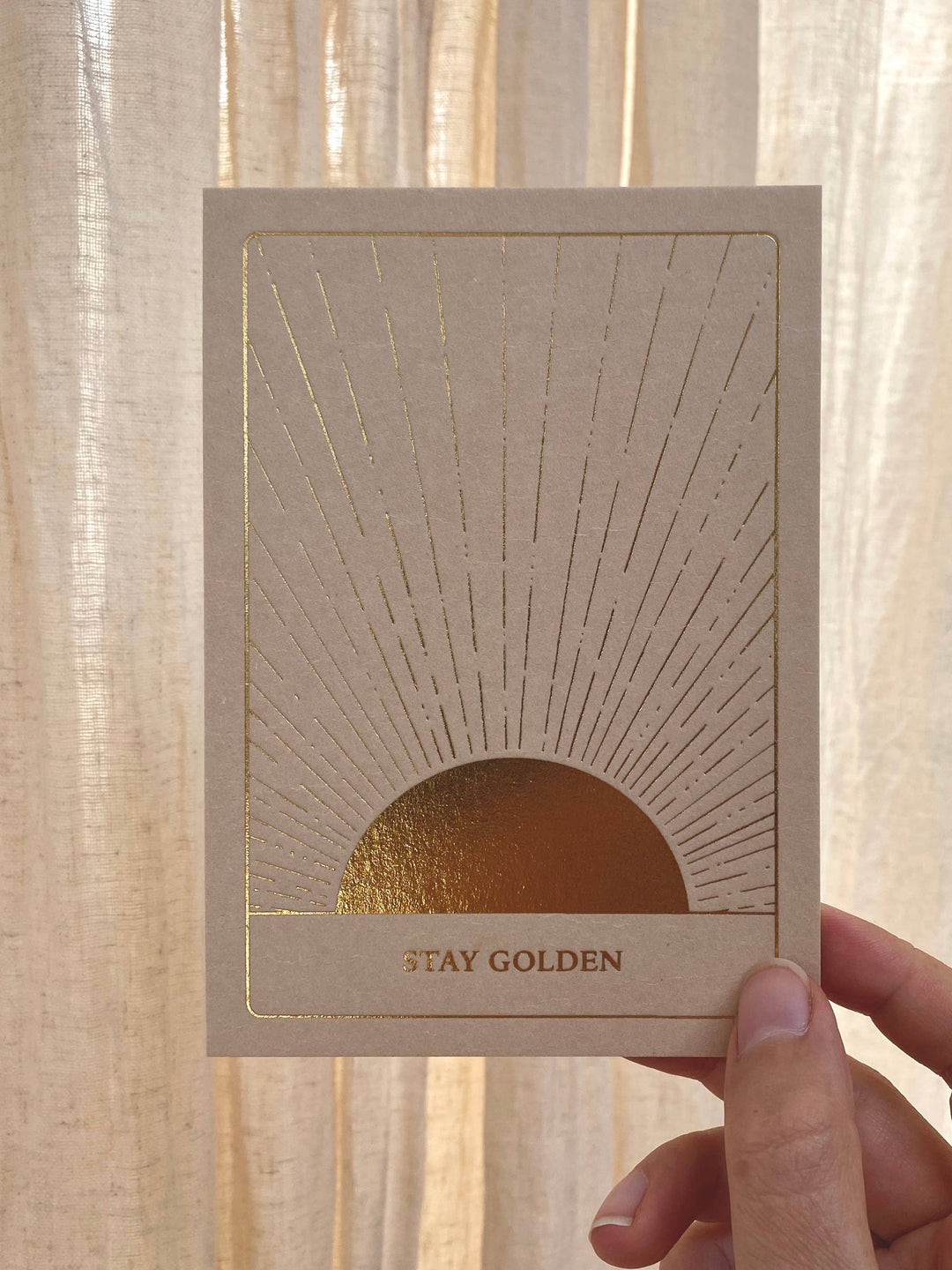 Card with sunburst design and 'Stay Golden' text held against a light curtain background