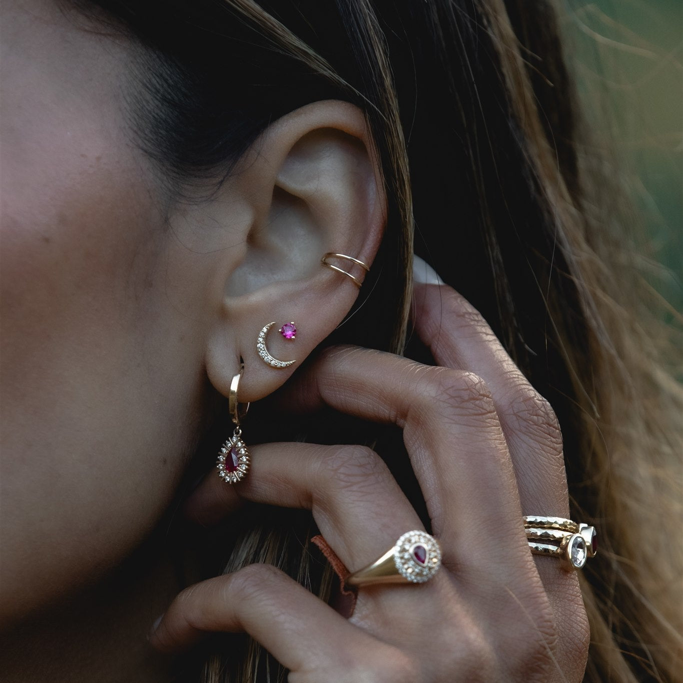 Close-up of an ear with gold hoop earrings and a hand touching the ear, wearing additional jewelry.