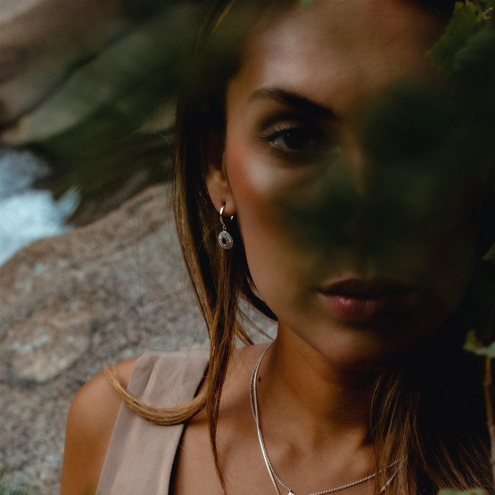 Woman with earrings and a necklace standing outdoors with greenery in the background