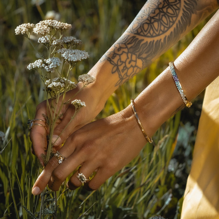 Person holding a bundle of flowers with tattoos and jewelry in a natural setting