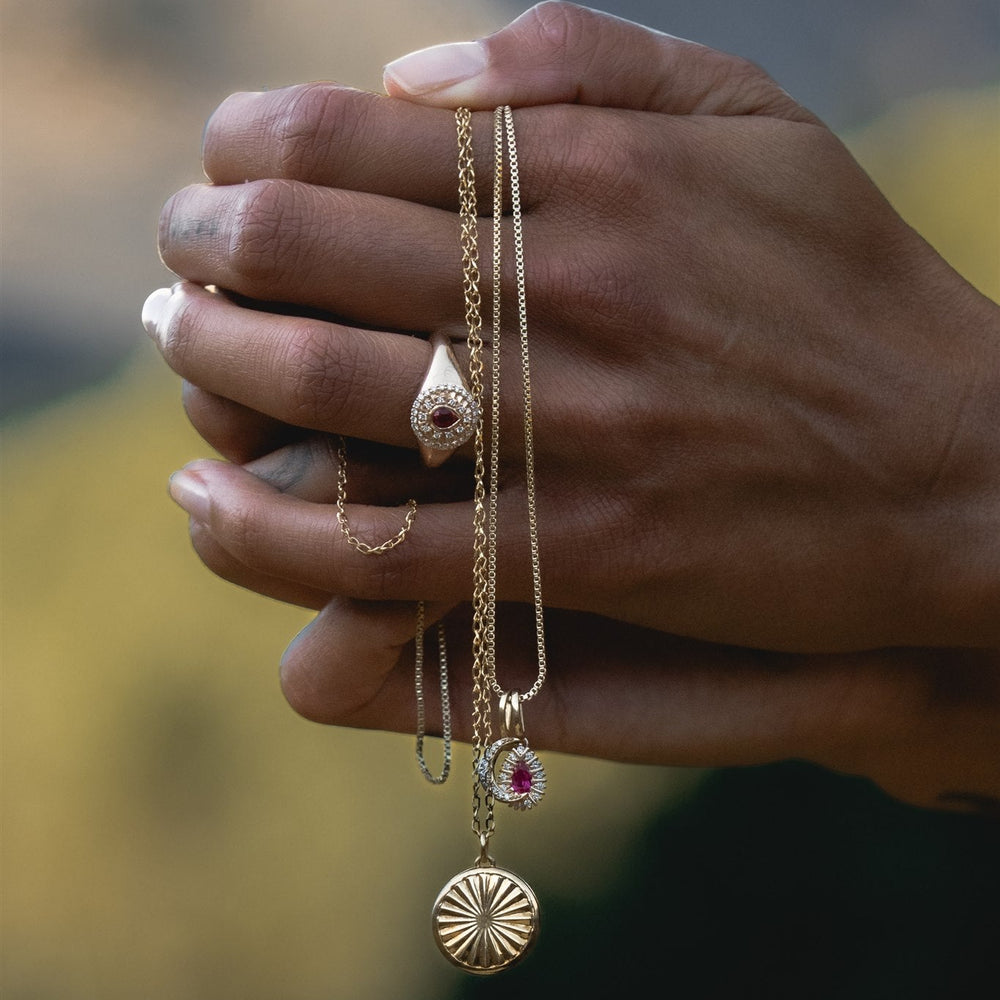 Hand holding three gold necklaces with various pendants against a blurred natural background