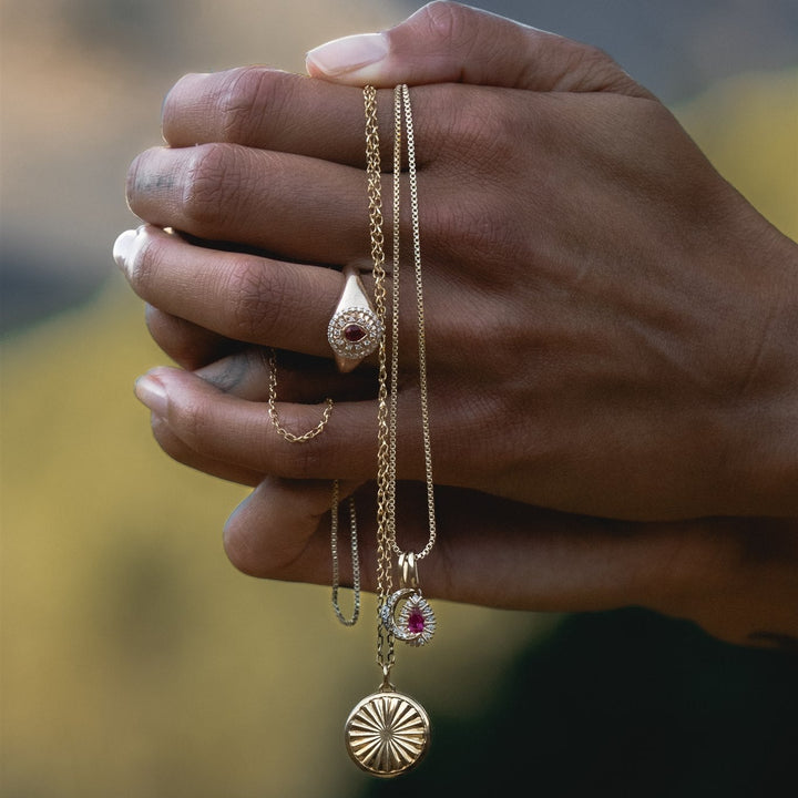 Hand holding three gold necklaces with various pendants against a blurred natural background