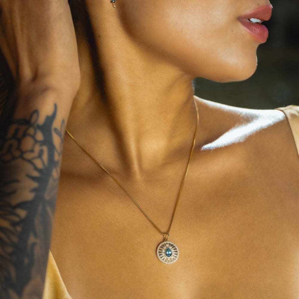Close-up of a woman wearing a gold necklace with a pendant, with a blurred background.