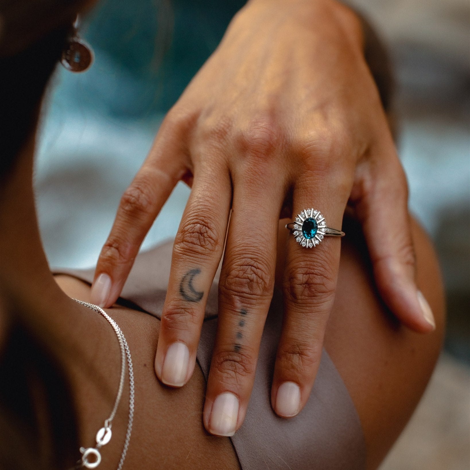 Close-up of a hand wearing a silver ring with a blue gemstone, with tattoos on the fingers.