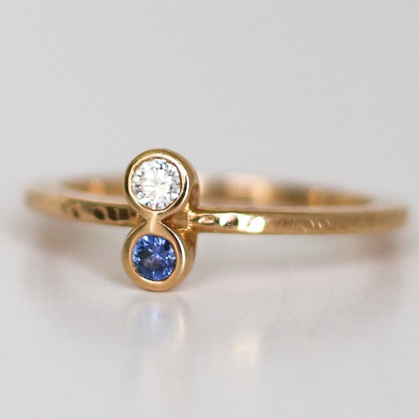 A stack of two rings, one with a round blue Montana Yogo Sapphire and the other with a round clear diamond, set in 14K gold with a hammered finish.