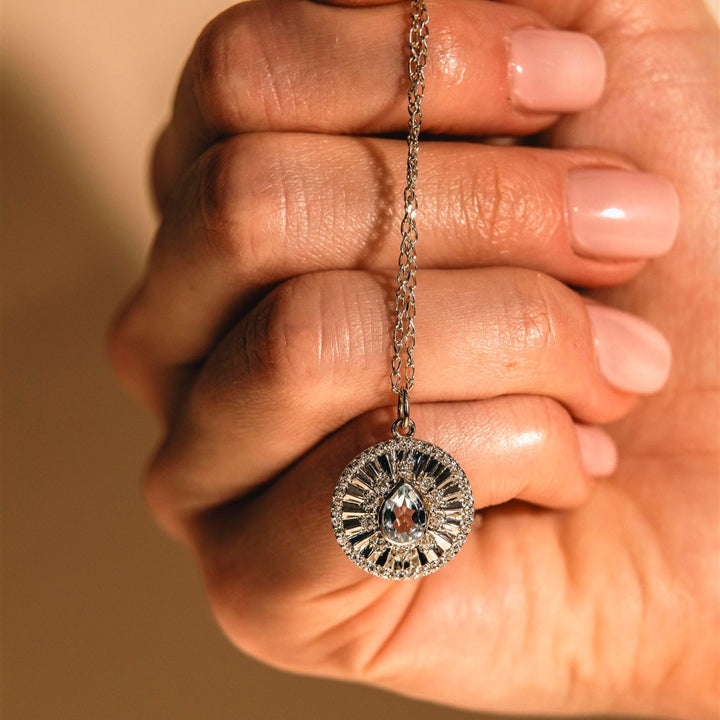 Hand holding a silver necklace with a decorative pendant against a beige background