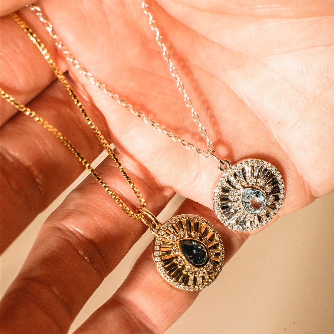 Two gold necklaces with intricate pendants held by a hand against a soft pink background.