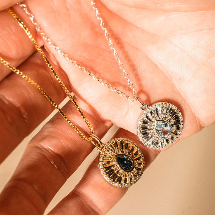 Two gold necklaces with intricate pendants held by a hand against a soft pink background.