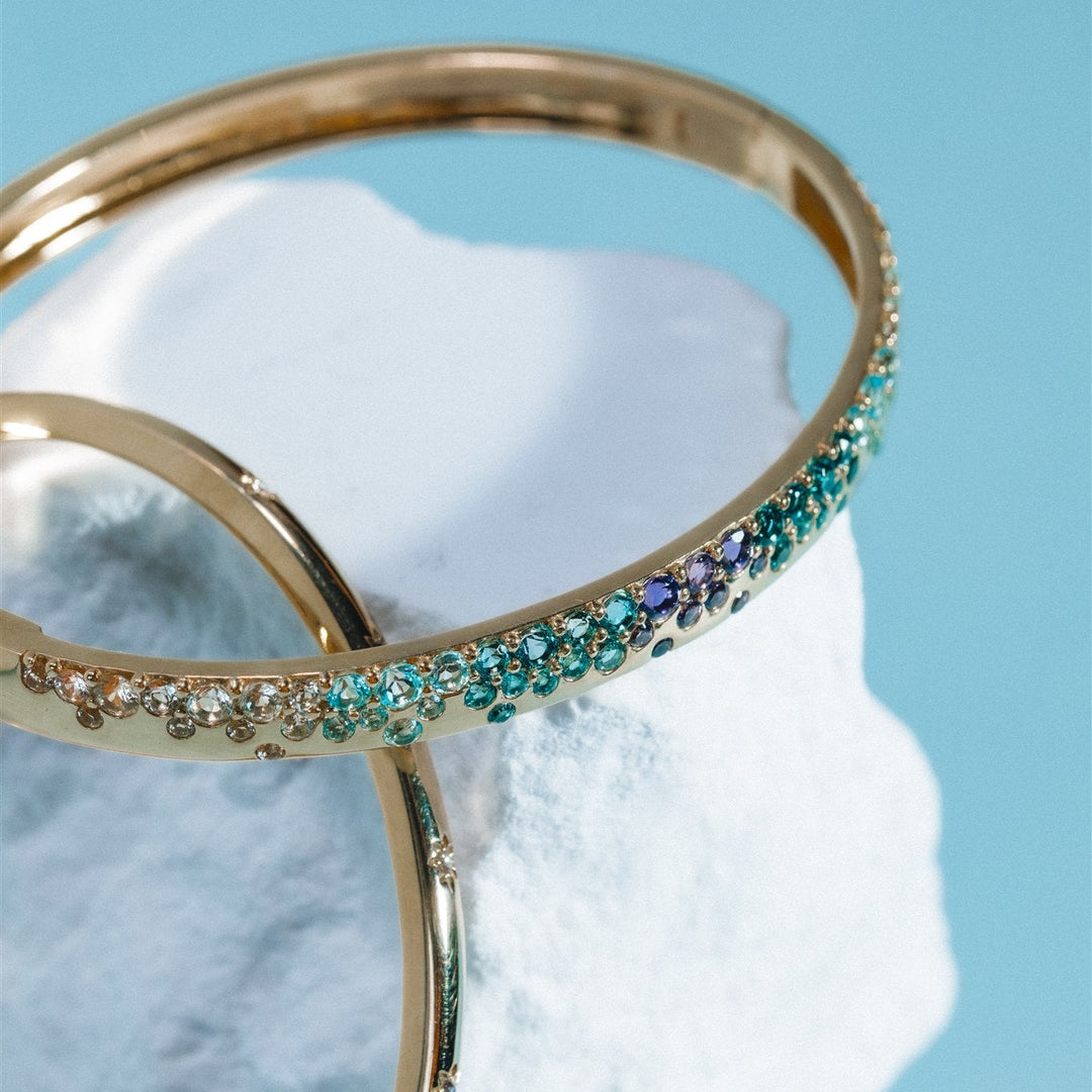 Gold bangles with gemstones on a textured white surface with a light blue background
