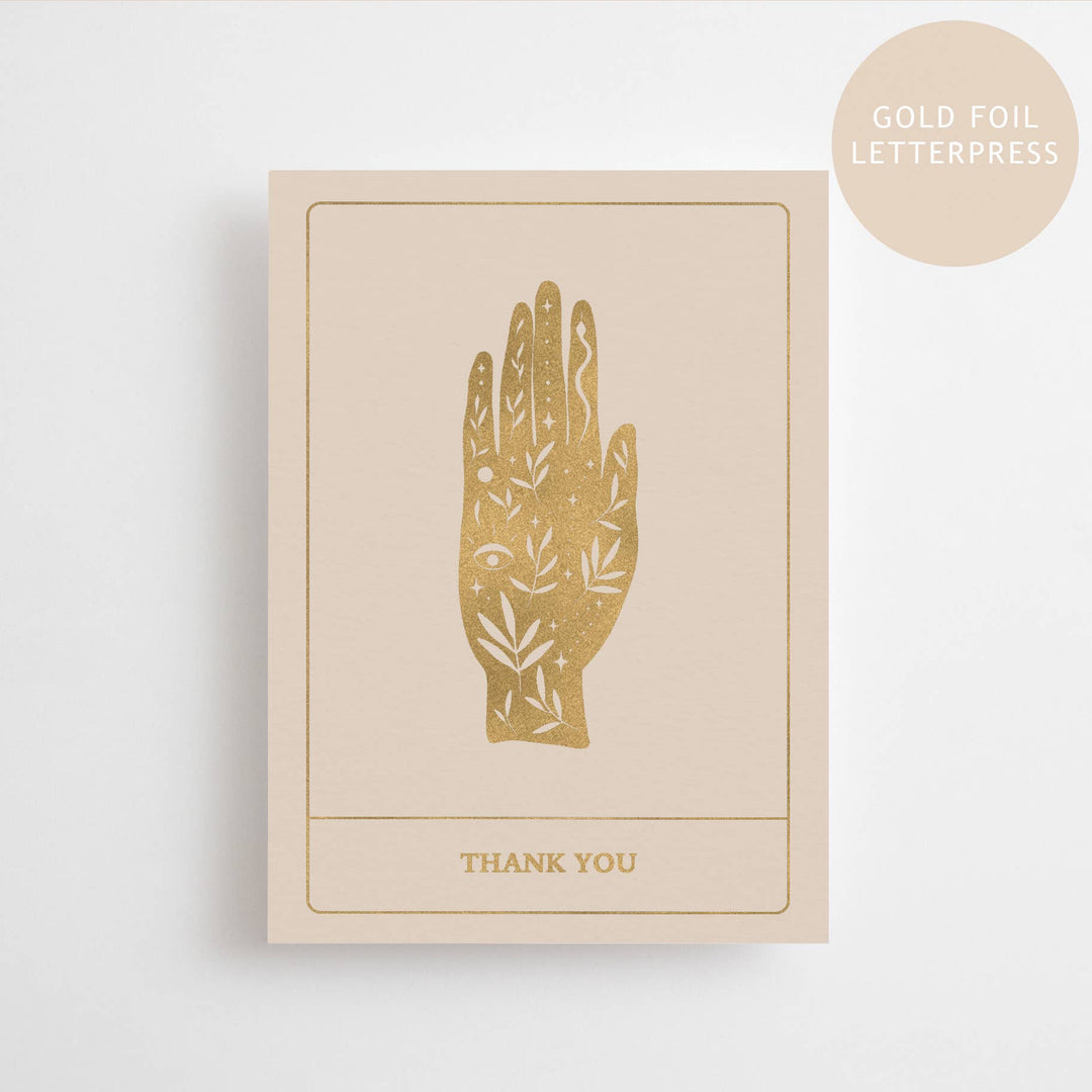 A beige colored postcard with a gold foil printed design of a hand and the words 'THANK YOU' below it, displayed on a neutral background.