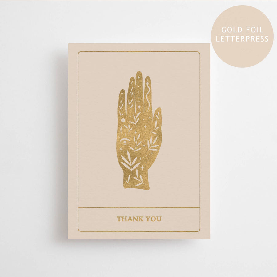 A beige colored postcard with a gold foil printed design of a hand and the words 'THANK YOU' below it, displayed on a neutral background.