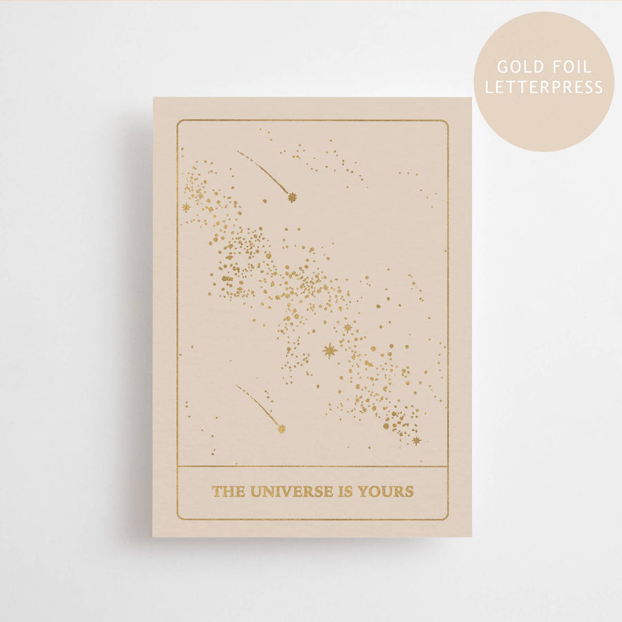 A beige postcard with gold foil letterpress text and designs, featuring the phrase 'THE UNIVERSE IS YOURS'.